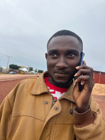 A medium shot captures a Black man in his late 20s or early 30s holding a dark-colored smartphone to his right ear and speaking, with his gaze directed upwards and to his left. He is wearing a brown, collared jacket over a red and white patterned shirt. The jacket is zipped up, and a metal button is visible on the left lapel. The man has short, dark hair and a short beard.
The background suggests an outdoor, rural or semi-urban setting. A dirt road is prominent in the foreground and extends into the middle ground. To the left of the road, there are several buildings, some appearing to be small shops or homes with corrugated metal roofs, as well as a utility pole and some trees. A dark-colored car is parked on the side of the road. To the right of the man, there is a large, red structure, possibly a building or wall, and a pile of what appears to be hay or straw. The sky is overcast with light gray clouds. The overall impression is a scene in the city of Durumi, Nigeria, possibly on the outskirts or in a less developed area.