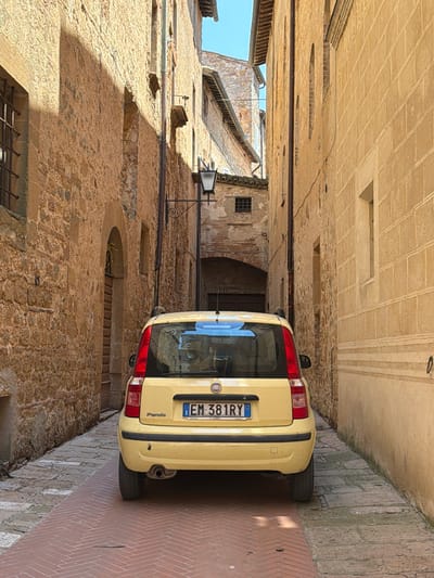 The image shows a narrow cobblestone alleyway in Pienza, Italy, with a yellow Fiat Panda parked in the middle. The buildings on either side of the alley are constructed of stone and plaster. The Fiat Panda is facing away from the camera, and its license plate reads "EM 381RY". The alley is well-lit by sunlight, and the sky is clear and blue. The stone buildings have arched doorways and small windows, typical of historic Italian architecture. The cobblestone paving is laid in a herringbone pattern in the foreground, transitioning to a more irregular pattern further down the alley. There are no people visible in the image.