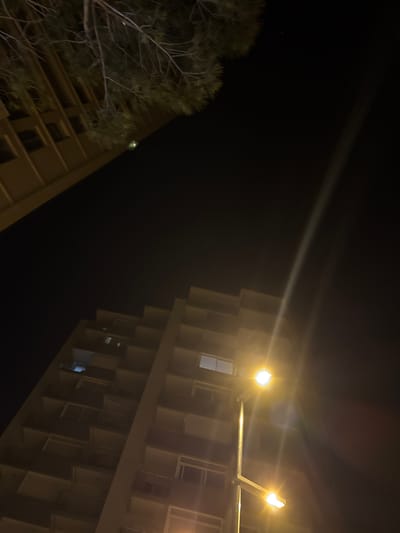 The image is an angled, low-light nighttime shot taken from below, looking up at two apartment buildings and a street lamp. The building on the left, partially visible, is a light brown color with visible windows and balconies. The larger building dominating the right side of the frame is also light brown, featuring a repetitive pattern of balconies and windows. A modern street lamp with two illuminated bulbs stands in the foreground, casting a bright yellow light that creates lens flares and streaks across the dark sky. A tree branch with pine needles hangs from the upper left corner, its silhouette dark against the night. The scene appears to be urban, with the characteristic architecture of apartment blocks and street lighting. The location is identified as Platja D'aro, Spain. There are no people visible in the frame, and no discernible actions or interactions are occurring. The environment is dark, suggesting nighttime, with artificial lighting from the street lamp and some windows.