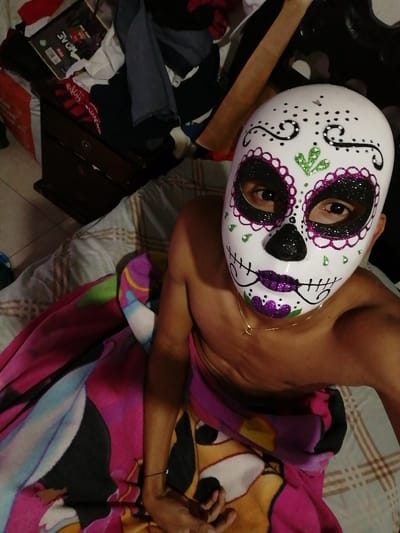 A shirtless person wearing a sugar skull mask is partially covered by a pink blanket with a cartoon character on it. The person is sitting on a striped bed. In the background is a dark wooden dresser with items piled on top, including clothes and a box. The mask is white with purple and green glitter details. The person's eyes are visible through the mask's eye holes.