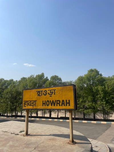 The image depicts a yellow signpost with the word "HOWRAH" written in English and Hindi. The signpost is located on a concrete surface near a tree-lined road, under a clear blue sky. It marks the location of Howrah, a city near Anajpur, India.