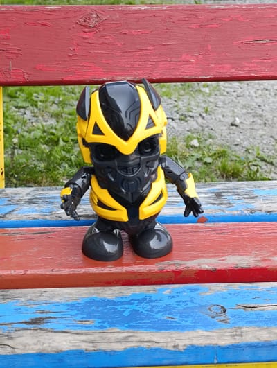 A toy robot, resembling Bumblebee from the Transformers franchise, stands on a weathered, multi-colored wooden bench. The robot is predominantly yellow and black, with intricate detailing on its head and body, and articulated arms. The bench has red, blue, and gray painted planks, showing signs of wear and peeling paint. Behind the bench, a patch of green grass and gravelly ground is visible. The background is out of focus, suggesting an outdoor setting. The image is taken from a slightly elevated perspective, looking down at the robot. The scene appears to be in Bogotol, Russia.