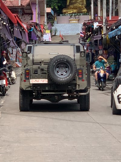 In a street scene set in Mae Sai District, Thailand, a beige SUV with a spare tire mounted on its rear is parked. The SUV has a license plate with Thai characters, which are white text on a red background. To the left of the SUV, a portion of a large golden Buddha statue is visible, suggesting the proximity of a temple or religious site.
In the background, a man in a turquoise shirt is riding a dark-colored scooter. He is touching his head, possibly indicating a moment of reflection or discomfort. The street is lined with buildings and stalls, some with colorful awnings and signs, creating a bustling market atmosphere. One building has a large white facade and what appears to be a flag with red and white stripes, possibly the Thai flag.
Several motorcycles are parked near the rear of the SUV, and one appears to be a red and white model with visible text on its license plate and fairing. The scene is illuminated by natural light, suggesting it is daytime. The overall impression is one of a typical day in a lively Thai district, with a mix of vehicles, local architecture, and a hint of the region's cultural identity.