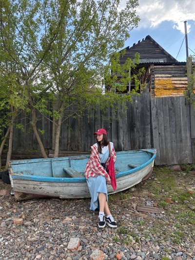 A young woman wearing a red cap, a white t-shirt, a denim dress, and a red and white plaid shirt is sitting in a light-blue boat. The boat is partially nestled against a weathered, wooden building with a yellow section. A tree with green leaves stands to the left of the building and boat. The ground surrounding the boat is covered in small rocks and some patches of grass. The background reveals a blue sky with clouds. This image was taken in the city of Овсянка, Russia.