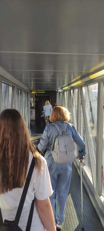 The image captures an interior view of an airport jet bridge in Khrabrovo, Russia, looking towards the terminal. Three individuals are visible walking through the bridge. In the foreground, the back of a person with long, brown hair is seen, wearing a white t-shirt and a black backpack strap. Ahead of them, a woman with short, blonde hair is walking away from the camera. She is wearing a light blue, long-sleeved outfit and carrying a light grey backpack. To her right, a person is partially visible, using a silver walking cane. Further down the jet bridge, a figure in a dark uniform, likely an airport employee, stands near an open doorway. The jet bridge features a metallic ceiling with fluorescent lighting and large windows along the right side, revealing an exterior view with sunlight. The floor of the jet bridge is covered with a textured dark grey material.
