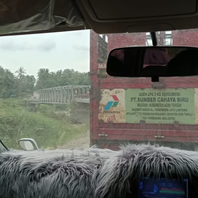The image, taken from inside a vehicle, shows a view of a road with a bridge in the distance and a red truck on the right. The truck has a sign that says "AGEN LPG 3 KG BERSUBSIDI PT. SUMBER CAHAYA BIRU WILAYAH: KABUPATEN ACEH TENGAH". The environment appears to be rural with lush green vegetation and trees. The sky is overcast, suggesting it might be raining or the weather is generally gloomy. The foreground of the image is dominated by a fuzzy grey material, likely a car accessory, and a screen displaying what appears to be audio level meters. The bridge in the background is a truss bridge made of metal. There are no discernible people clearly visible in the scene. The overall impression is a snapshot of a journey through a somewhat remote area in Indonesia.
