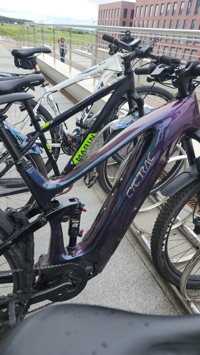 This media file shows a close-up of several bicycles parked outdoors. The foreground is dominated by a purple, iridescent bicycle frame with "CYCTRAC" written in silver lettering. The bicycle has a suspension shock absorber visible. To the left and slightly behind it is a white bicycle frame, partially obscured. Further back, other bicycle wheels and frames are visible, some with visible tire brands like "MAXXIS". The bicycles are parked in a row, likely in a bike rack, on a paved surface with a textured pattern. In the background, a brick building with many windows is visible, suggesting an urban or institutional setting. There is also a glimpse of greenery and a person in the distance walking. The lighting suggests it is daytime. The scene appears to be set at the Skolkovo Innovation Center in Russia.