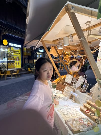 A young East Asian woman with dark hair pulled back is standing at a market stall in Yuzhong District, China. She is wearing a light pink t-shirt with a graphic design. The stall is wooden and displays various handcrafted jewelry, including necklaces, bracelets, and earrings, arranged on a white lace tablecloth and wooden stands. A round mirror with a decorative frame hangs on the stall, reflecting the woman's profile. In the background, a building with traditional Chinese architecture is visible, along with a brightly lit shop displaying drinks and seating areas. The street is paved, and the scene appears to be at dusk or night, with artificial lighting illuminating the stall and surrounding area.