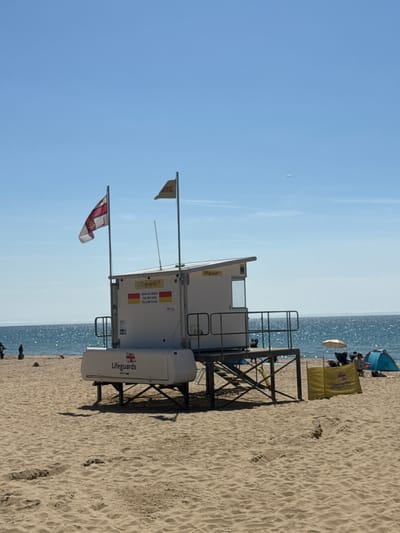 This is a photograph of a lifeguard station on Bournemouth Beach in Bournemouth, United Kingdom. The station is a white, elevated structure with a roof and an enclosed cabin. There are two flags flying from poles next to the station: one is a red and white ensign, and the other is a yellow flag with a red cross. Text on the side of the station reads "Lifeguards" and "SWIM BETWEEN THE RED AND YELLOW FLAGS." A yellow windbreak with "Lifeguards" printed on it is positioned in front of the station. The beach is made of sand, and the sea is visible in the background. Several people are present on the beach, some sitting under umbrellas and others walking. There are also a couple of beach tents visible. The sky is clear and blue.