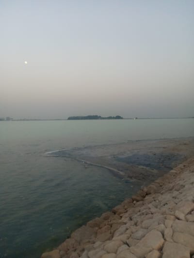 The image depicts a coastal scene at dusk in Al Khobar, Saudi Arabia. A body of water, possibly the Arabian Gulf, stretches towards the horizon under a pale, overcast sky with a faint moon visible. A small, vegetated island is visible in the distance. The shoreline is reinforced with large, light-colored rocks. The water's edge meets a sandy or muddy transition zone. There are no people visible in the scene.