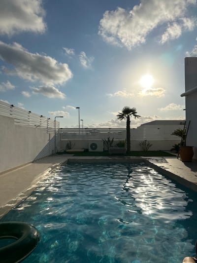 This image depicts a private rooftop swimming pool area in the city of Benijófar, Spain. The sun is shining brightly in a partly cloudy sky, casting reflections on the rippling blue water of the pool. A black inflatable ring is partially submerged in the foreground. Beyond the pool, there is a paved area with a white slatted fence and a low white wall. In the background, a small palm tree stands tall, surrounded by some plants and an outdoor air conditioning unit. A ladder leans against a large terracotta pot on the right side of the frame. The architecture suggests a modern residential building. The scene is set during daytime, likely late afternoon given the angle of the sun.