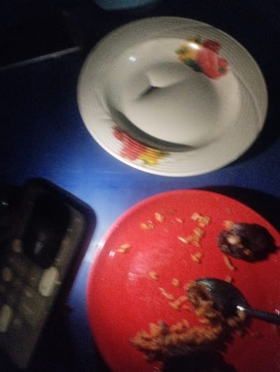 An overhead, low-light image captures a dining surface, presumably in the city of "Healtcare," Nigeria. The specific location within the city is not discernible. The scene features two ceramic plates and a mobile phone on a dark blue or black table surface. In the upper right, a clean, white plate with a floral pattern of red and yellow flowers along its rim is present. In the lower center and right, a red plate contains remnants of a meal, consisting of rice, a dark solid food item (possibly meat or a date), and a metallic spoon resting within the food. To the left of the red plate, a dark-colored mobile phone, possibly enclosed in a black case, is visible. No individuals are present. The items suggest a recently concluded or ongoing meal.