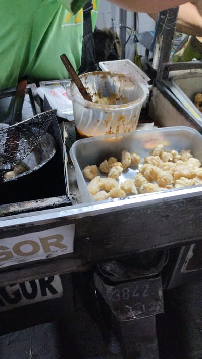 This is a medium-angle, eye-level shot of a food stall, likely in Sukodono, Indonesia, featuring the preparation of fried food. In the foreground, a metal container holds a pile of golden-brown, puffy fried snacks, possibly pork rinds or some form of crispy dough. To the left, a deep fryer with a black enamel finish contains a metal mesh basket submerged in hot oil, with several pieces of food being fried. A wooden utensil rests in a clear plastic container holding a brownish, chunky sauce or dip to the right of the fryer. The background shows a person wearing a bright green t-shirt and black straps, suggesting they are the vendor. The stall appears to be outdoors, possibly at a market or street side, with hints of other stalls or structures in the blurred background. Signs with text are partially visible, including "GOR" and "GUR", and a number "3842" is stamped onto a metal component of the stall. The overall scene conveys a sense of active food preparation and sale.