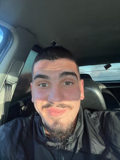 The image presents a close-up, head-and-shoulders portrait of a young adult male situated within a vehicle in the city of Kovachitsa, Bulgaria. The subject, positioned centrally and facing the camera directly, has short dark hair, dark eyebrows, light brown eyes, a mustache, and a short beard. He is wearing a dark, zippered jacket and exhibits a subtle smile. Strong natural light from the left illuminates the left side of his face and upper torso, creating distinct shadows on the right. The vehicle's interior features a gray headliner, a visible seatbelt mechanism on the left, and a rear window through which a blurred exterior environment is discernible. A small, dark, rectangular object hangs near the top right quadrant of the rear window.