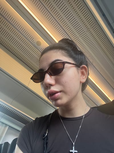 A young woman with dark hair pulled back in a bun, wearing sunglasses and a dark brown t-shirt, is positioned in the foreground of the image. She is looking towards the camera, and her face is partially obscured by the sunglasses. A silver necklace with a cross pendant is visible on her chest. The background features a textured ceiling with recessed lighting, suggesting an indoor environment, possibly a train carriage or a similar mode of transport. The lighting appears to be artificial, casting a warm glow. The image does not provide any specific information or visual cues to determine a location in Vejprnice, Czechia.