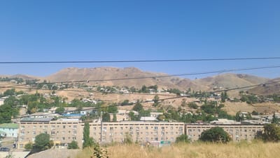 The image shows a wide shot of a residential area in the city of Sülüktü, Kyrgyzstan, under a clear blue sky. In the foreground, dry, golden grass and shrubs partially obscure the view of several buildings. Dominating the mid-ground are several multi-story apartment buildings, characterized by their beige and grey facades, repetitive window patterns, and flat roofs. Some of these buildings appear to be of socialist-era architecture. Scattered amongst the apartment blocks are smaller houses, many with pitched roofs, and a variety of trees, predominantly green and leafy, suggesting a temperate climate. The background features rolling hills and mountains, barren and arid in appearance, with houses and structures sparsely dotting their slopes. Several power lines crisscross the scene, connecting different parts of the city and indicating infrastructure. The overall impression is of a semi-arid, mountainous urban environment.