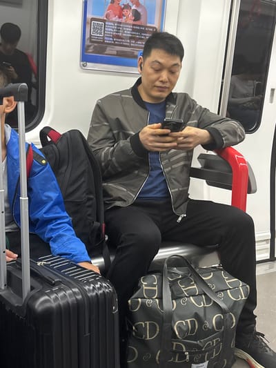 The scene depicts the interior of a subway car in Chancheng District, China. An adult male, approximately 30-40 years old, with short dark hair, is seated on a metallic grey bench. He is wearing a grey bomber jacket with horizontal grey stripes, a blue t-shirt, black pants, and dark athletic shoes with white and red soles. He has a small black earbud in his right ear and is holding a black smartphone with both hands, looking down at its screen. A black backpack with red accents rests on the seat to his left. On the floor in front of him, between his feet, is a black duffel bag patterned with repeating light gold "CD" logos. To the immediate left, partially visible, is a person wearing a bright blue jacket with a yellow accent, seated next to a black hardshell rolling suitcase with a silver metallic extendable handle. The subway car features white interior walls, a red armrest for the main subject, and a framed poster with a family image, a QR code, and Chinese text above the seated man. Reflections of other passengers and the train interior are visible in the windows and doors on both sides of the car.