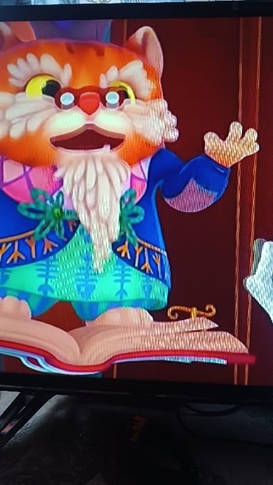 The image displays a photograph of a screen exhibiting an animated scene. The primary subject is a cartoon-style anthropomorphic cat with orange and white fur, yellow eyes, a red nose, and circular spectacles with light blue lenses. It possesses a prominent white mustache and beard, and its mouth is open. The cat is clad in a multi-colored robe: blue predominantly, with pink and purple checkered patterns on the upper chest, and a teal-green section at the waist featuring geometric designs. Two green bow-like elements are affixed to its chest. The cat's right paw is raised, fingers extended. In front of the character, an open book with light-colored pages and a red spine is visible, with a small, golden, ornate object on its right page. The background is a solid reddish-brown, indicative of a curtain or wall. The scene is located in Kasansay, Uzbekistan.