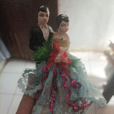 This image captures a decorative figurine set, likely a wedding cake topper, held by a human hand in an indoor environment within Pasir Muncang, Indonesia. The set features two painted figures: a male figure in a black suit positioned slightly behind a female figure in a white and gold strapless garment. Both figures possess dark hair and simply rendered facial features. Green plastic foliage and a red fabric bow adorned with smaller pink/white flowers are situated centrally between them, resting on a base covered by light blue lace from which multiple dark pink ribbons cascade. The human hand supports the base from the lower left. The blurred background shows a dark, textured surface on the left and a prominent, overexposed light source on the right. The image exhibits visible digital noise throughout.