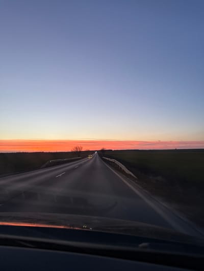 The image depicts a rural road at sunset or sunrise, viewed from inside a vehicle. The sky displays a clear gradient: intense orange and red hues dominate the horizon, transitioning upwards through lighter orange and pink bands to a pale blue, then a deeper blue at the top. A dark, asphalt two-lane road stretches into the distance, marked by white dashed lines. Metallic guardrails line both sides of the road. Two sets of headlights are visible in the far distance, indicating approaching vehicles. Flat, dark agricultural fields extend to the left and right beyond the guardrails, meeting a distant tree line composed of sparse, bare deciduous trees or bushes silhouetted against the horizon. The very bottom of the image shows a dark, reflective surface consistent with a vehicle's dashboard or windshield interior. The scene is located on a rural road in or near Vaglen, Bulgaria.