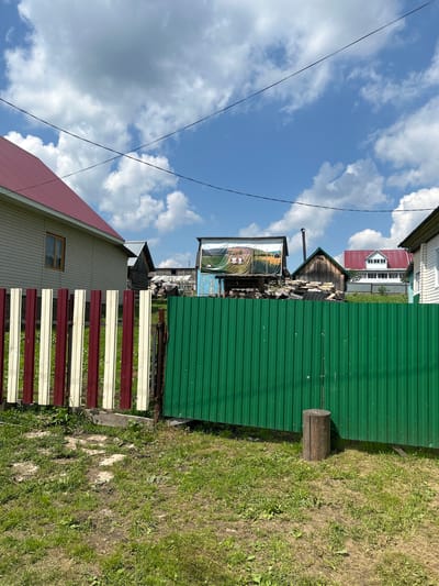 The image depicts a rural scene in Novomullakaevo, Russia, under a bright blue sky with scattered white clouds. In the foreground, a striped fence made of alternating red and cream vertical wooden planks is visible on the left, partially obscured by a green corrugated metal fence on the right. A wooden log stands upright in front of the green fence. Behind the fences, several structures are present. To the left, a light-colored house with a red metal roof and a single window is seen. Further back, various wooden buildings with different roof styles and materials are scattered across the landscape. One structure has a large banner or tarp displaying an image of cows in a field. The ground is covered in patchy green grass and dirt. Power lines are visible stretching across the sky.