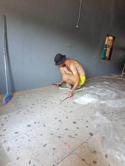 A shirtless man wearing a bucket hat and yellow shorts is crouching on a floor covered in terrazzo tiles. He is holding a ruler and what appears to be a marker or pencil, suggesting he is measuring or marking the tiles. To his right, plastic sheeting covers part of the floor and extends into the background. To his left, a broom with a blue brush stands upright. The scene appears to be indoors, possibly a construction or renovation site, with grey walls. A small, recessed shelf in the wall behind the man contains a teal-colored bottle and some electrical components. The lighting suggests it is daytime.