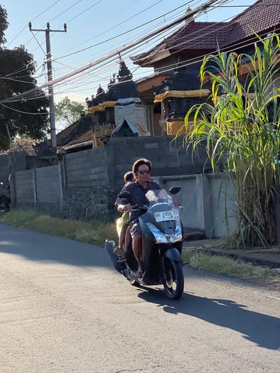 The image depicts two people riding on a scooter along an asphalt road in Kubu, Indonesia. The person in the front appears to be a man, and the person behind him is likely a woman. They are both wearing casual clothing. The scooter has a license plate visible with the text "DK 2858 TF 07.28".
The scene is set in a tropical environment, with lush green vegetation growing on the side of the road. There are also buildings with traditional Indonesian architecture visible in the background, including a structure with a red tiled roof and intricate carvings. Electrical wires are strung across the scene, supported by utility poles, suggesting a residential or semi-rural area.
The time of day appears to be late afternoon or early evening, as indicated by the long shadows cast on the road. The sun is low in the sky, casting a warm glow on the scene. The weather seems to be clear and sunny.
The overall mood of the image is relaxed and everyday, capturing a common mode of transportation and a glimpse into daily life in Indonesia. The focus is on the journey and the people, with the surroundings providing context for their location and activity.