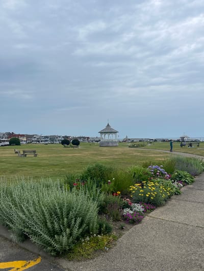 This is a vertical, eye-level, full shot of a park-like setting by the ocean. The foreground features a paved walkway and a vibrant flower bed filled with a variety of colorful flowers, including yellow, purple, white, and red blooms, as well as lush green foliage. Beyond the flower bed, a large expanse of neatly mowed green grass stretches into the distance.
In the middle ground, a white gazebo with an ornate roof stands prominently on the lawn. Further back, a line of houses with red-tiled roofs and white facades forms a picturesque coastal village. Beyond the houses, a body of water is visible, with a few boats docked and a large ship. Several people are scattered throughout the scene, some walking along the path, others standing near the water, and one person is taking a picture.
The sky is overcast with a pale, muted blue color, suggesting a cloudy or overcast day. The overall impression is one of a tranquil and scenic coastal town, possibly during the daytime. The well-maintained park, blooming flowers, and charming architecture contribute to a peaceful and inviting atmosphere. The yellow painted line on the edge of the walkway adds a touch of detail to the scene.