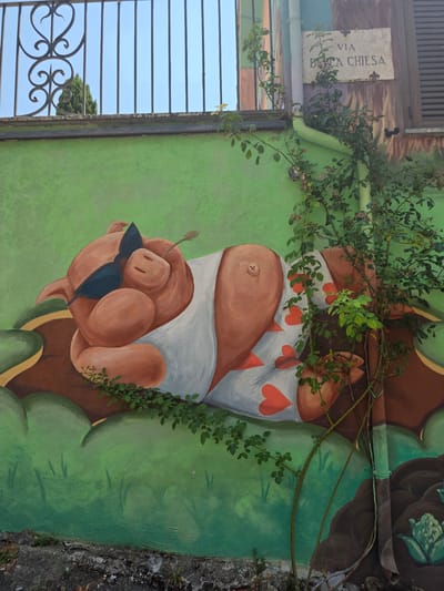 This image showcases a vibrant mural painted on a wall, likely in Viterbo, Italy, as suggested by the "VIA B.A. CHIESA" street sign visible. The mural depicts a whimsical character, a pig with a human-like body, wearing a white garment adorned with red hearts and a dark blue bow tie. The pig has rosy pink skin and a small tail. It appears to be leaning or resting, with a gentle expression.
The background of the mural is a bright, textured green, possibly representing foliage or a grassy landscape. Intertwined with the mural and extending from the top of the image is a living vine with lush green leaves, adding a natural and dynamic element to the artwork. A rusty downspout runs horizontally across the mural, further integrating the art with the building's structure.
To the left of the mural, a section of a dark, slatted window or grate is visible, revealing a clear blue sky and a distant green tree. The presence of the street sign and the architectural style hints at an urban or semi-urban setting, possibly along a street or in a courtyard. The overall tone of the mural is playful and cheerful, with the bright colors and the charming subject matter creating a sense of lightheartedness. The time of day appears to be daytime, given the bright sunlight casting subtle shadows.
