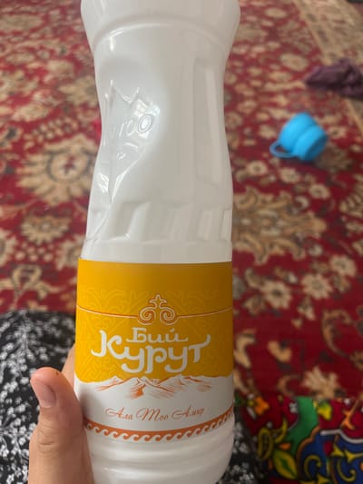 A person's hand holds the bottom portion of a white plastic bottle with an orange label. The label features decorative elements, mountains, and text in a script that appears to be Cyrillic and Latin. The background is a red carpet with a floral pattern. A blue plastic cup is visible in the blurred background on the carpet. The scene appears to be indoors. The location is the city of Ташлак, Kyrgyzstan.