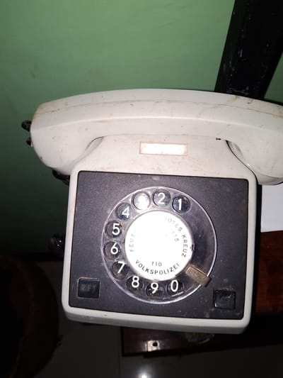 An antique white rotary dial telephone is positioned on a dark wooden surface in what appears to be a dimly lit indoor setting. The telephone is dusty and shows signs of age. The dial features numbers from 0 to 9, and text indicating "FEUERWEHR" and "VOLKSPOLIZEI". A black handset is attached to the telephone. In the background, a green wall is visible, with a black cylindrical object to the right. The location is specified as Sendangrejo, Indonesia.