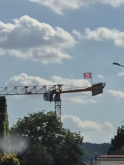 A construction crane is visible against a blue sky with scattered white clouds. A small red flag with a white symbol is attached to the crane. The crane is yellow and gray, with a cabin at the end of the horizontal arm. Green trees and foliage are in the foreground, with a hint of a building's roof at the bottom of the frame. The background shows a tree-covered hillside. The scene appears to be in a semi-urban or suburban area.