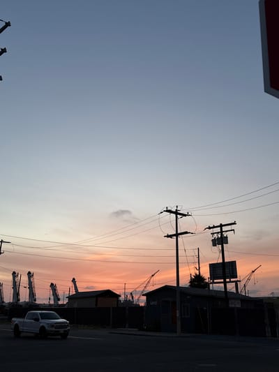 The image shows a view of National City, California, United States of America at what appears to be sunset or sunrise. The sky transitions from a pale blue-gray to a gradient of pink and orange near the horizon. Several utility poles with power lines are visible in silhouette against the sky. In the lower right, the image shows a portion of a street, a building (possibly an industrial structure or storage facility), and a white pickup truck parked on the side of the road. Several cranes are visible as well, adding to an industrial port or manufacturing environment. The overall lighting suggests either early morning or late evening.