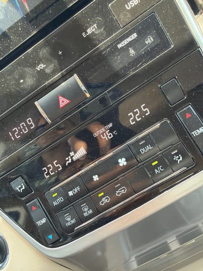 This media file captures a close-up, angled view of a dusty vehicle dashboard, specifically focusing on the climate control and infotainment system. The scene is set inside a car, likely in a hot climate, as indicated by the "OUTSIDE TEMP" display showing 46°C. The current time displayed is 12:09.
The central part of the image features two digital temperature readouts, both showing "22.5," suggesting the desired interior temperature setting. To the left, controls for volume ("VOL" with "+" and "-") and an "EJECT" button are visible, along with a "USB" port label. A hazard light button with a red triangle is prominent.
The right side of the image displays various climate control buttons: "DUAL" for dual-zone temperature control, "A/C" for air conditioning, "AUTO" mode, "OFF" for the climate system, and "TEMP" buttons (with red and blue arrows) for adjusting temperature. Icons for fan speed, air circulation, and front and rear defrosters are also present. A "PASSENGER" label is visible with an icon that typically indicates the status of the passenger airbag or seatbelt. The dashboard surfaces, particularly around the buttons and displays, are covered in a noticeable layer of dust. The interior color of the car appears to be a light beige or cream in the background.
