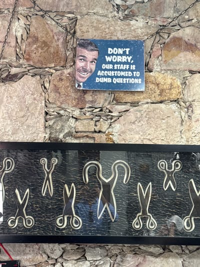 The image shows a stone wall on which a sign reading "DON'T WORRY, OUR STAFF IS ACCUSTOMED TO DUMB QUESTIONS" is displayed along with a picture of a smiling man, next to a framed display of decorative scissors. This likely exists in the city of San Miguel de Allende, Mexico, suggested by architectural features.