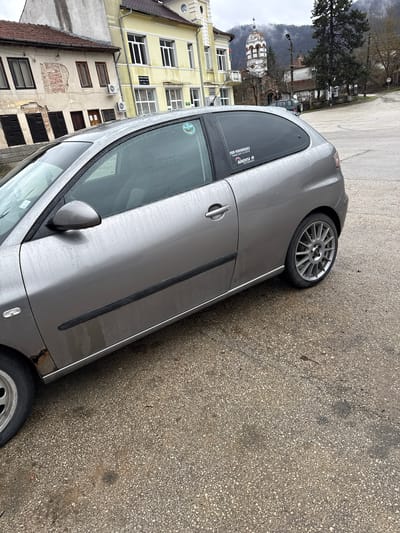 The image depicts a grey car parked on a wet asphalt road in what appears to be a city environment. Behind the car is a row of buildings of varying architectural styles. One building is a light yellow color, while others are older and show signs of wear. The dome of what seems to be a church is visible in the background. The image is taken on an overcast day, likely in Mikre, Bulgaria.