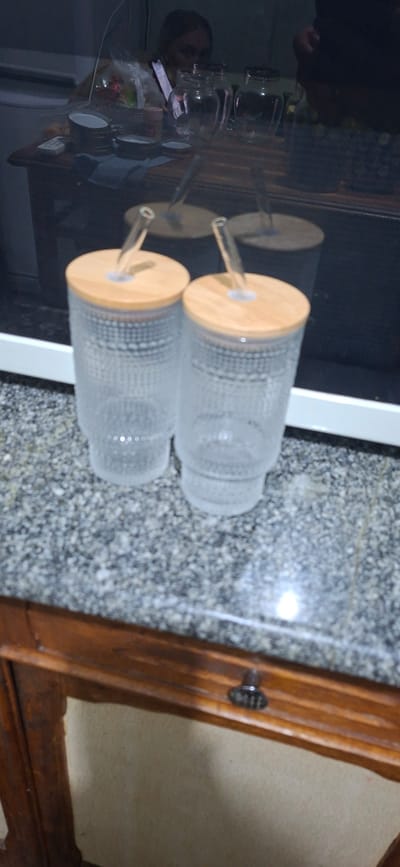 The image displays two clear glass tumblers with bamboo lids and glass straws. The tumblers have a textured pattern and are placed on a speckled countertop. In the background, a person is visible looking at a phone, with several glass jars and other items on a wooden surface, possibly a table. The overall scene appears to be indoors, in what seems to be a kitchen or dining area. There is no information within the image to suggest the location is in Shakhtarsk, Ukraine.
