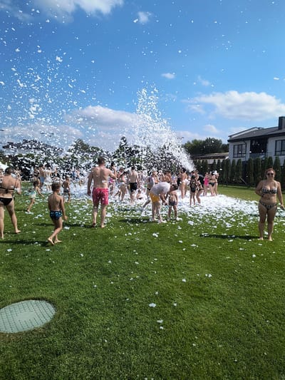 This media file shows a group of people enjoying a foam party on a bright, sunny day in Velyka Omeliana, Ukraine. The scene is set on a well-maintained green lawn, with a modern house visible in the background. The sky is a vibrant blue with scattered white clouds, indicating fair weather.
The primary activity is the foam party, with a foam machine spraying a large amount of white foam into the air and over the grass. A diverse group of people, dressed in swimwear, are scattered across the lawn, many interacting with the foam. Some are standing in the foam, while others are closer to the foam machine, which is not entirely visible but its output is apparent. The people appear to be enjoying themselves, with a generally positive and festive atmosphere.
Notable details include the bright daylight, suggesting it's likely daytime, possibly afternoon, given the activity. The presence of a modern house and manicured lawn suggests a residential or recreational area. The people are of various ages, including adults and what appear to be children. The foam itself is a prominent visual element, creating a playful and somewhat chaotic environment. There is no visible text in the media file. The overall impression is one of a joyful summer gathering.