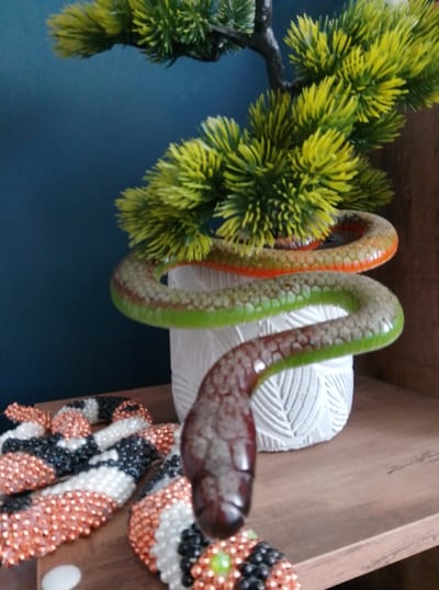 A coiled, plastic toy snake, predominantly green with orange and brown accents, is draped around a potted artificial plant. The plant, with bright yellow-green foliage, is housed in a white, textured pot. The snake's head is the closest object to the viewer, appearing dark brown and glossy. To the left of the snake and plant, on a wooden surface, are two intricately beaded objects that resemble coiled snakes or decorative pieces. These beaded items are made up of small, shiny beads in black, white, and a copper or rose-gold color. The background is a dark blue wall. The scene appears to be indoors, likely on a shelf or table, with a wooden surface visible in the foreground and on the right side of the image. The lighting suggests an indoor setting, possibly during the daytime, but there are no direct light sources visible. No people are present in the image, and no text is visible. The overall impression is of a decorative display.