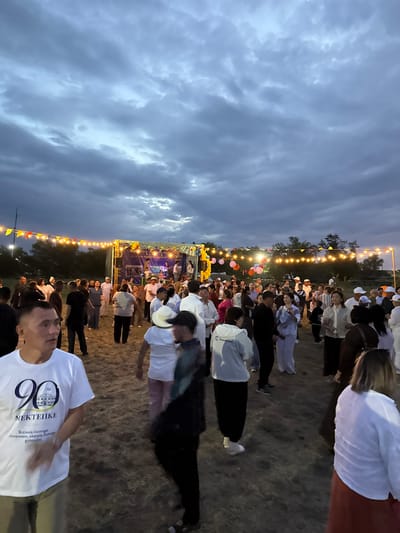 This media file depicts an outdoor evening gathering, likely a festival or celebration, in what appears to be a rural or semi-rural setting in Kazakhstan. A large group of people, dressed in a mix of casual and formal attire, are congregated in an open field. Many are wearing white t-shirts, some with visible logos or text.
The scene is illuminated by strings of warm-toned lights strung across the area, and in the background, a stage or platform is visible with more lighting and what seems to be a banner. There are also colorful balloons dispersed among the crowd. The time of day is dusk or early evening, with a cloudy sky overhead. The ground is covered in dry grass or dirt.
One person in the foreground is wearing a white t-shirt with the number "90" and text that appears to be in Cyrillic script: "90 MEKTENTEKE" and below it, "Бастауыш білімге, жоғары білімге, салтанатты, еңбекке, жастыққа." This text translates roughly to "Primary education, higher education, solemnity, labor, youth."
The overall atmosphere seems festive and communal, with people gathered together for an event. The cloudy sky suggests overcast conditions, but the lighting and the presence of many people indicate an active and lively occasion.