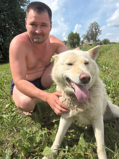 A shirtless man with short brown hair and stubble is crouching in a grassy field, petting a light-colored dog. The man is wearing dark blue shorts. The dog, a husky-mix with light fur, is lying down with its tongue hanging out and eyes partially closed. The background shows a green field with trees and a cloudy blue sky. The scene appears to be outdoors in a rural or park-like setting. The location is specified as Nizhnii Vishur, Russia.