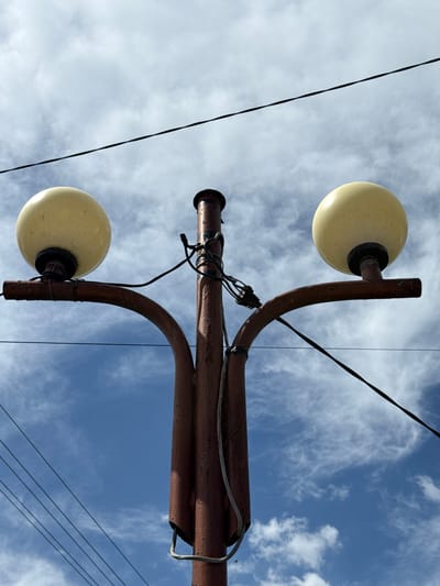 The image depicts a vintage street lamp in Ogradna, Bulgaria. The lamp consists of a rusted brown metal post with two curved arms. Each arm supports a milky yellow spherical light fixture. Electrical wires are haphazardly strung along the post and arms. The background is a bright blue sky with wispy white clouds. Power lines are visible in the periphery.