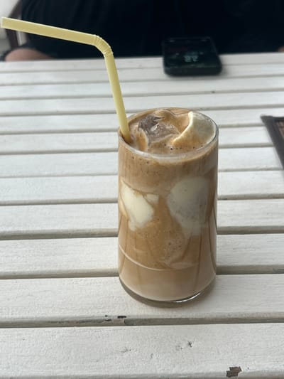 The image displays a tall, clear glass containing a layered iced coffee beverage situated on a white slatted table in the city of Dolni Tsibar, Bulgaria. The drink features a light brown liquid base, topped with a darker foamy coffee and several distinct white creamy components, along with multiple ice cubes. A yellow, flexible plastic straw is inserted into the beverage, bent near its top. The white table surface, composed of horizontal slats, shows minor wear. In the out-of-focus background, a person wearing dark clothing is partially visible to the upper left, while a dark-colored smartphone lies flat on the table to the upper right. A dark, textured object, possibly a placemat, is partially visible on the far right.