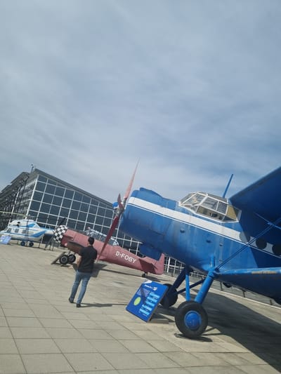 The image depicts two aircraft on display outside the Flugwelt Stuttgart aviation museum in Echterdingen, Germany. The primary aircraft is a blue and white Antonov An-2 biplane. Beneath it, a red and white aircraft is partially visible, with the registration "D-FOBY" painted on its fuselage. A person wearing a black shirt and jeans stands near the red and white aircraft, seemingly observing it. In the background, a modern glass and steel building is visible. The ground is paved with light-colored rectangular tiles. The sky is light blue with some clouds. A blue and white helicopter is visible in the distance. A blue sign is near the aircraft.