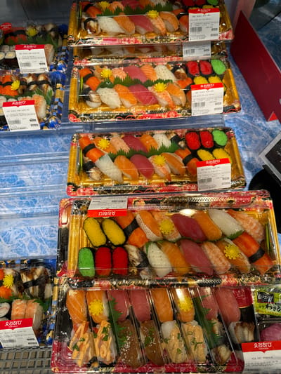 The image displays a close-up of several plastic containers filled with various types of sushi, neatly arranged on a shelf. The sushi includes nigiri, maki, and other varieties, with visible ingredients such as salmon, tuna, shrimp, eel, and rice. Some containers have labels in Korean and Vietnamese, indicating brands like "YORIHADA" and "LOTTE MART." The background shows a blue, textured surface, likely a refrigerator shelf in a grocery store. The lighting suggests an indoor retail environment. The scene is static, focusing solely on the product display. The city of Phu Thuy, Vietnam is not visible or implied in the image.