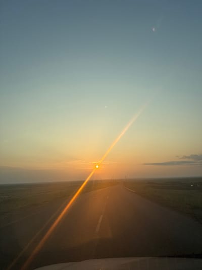The image depicts a vast, open landscape, likely in Berdyanka, Russia, during what appears to be sunrise or sunset. The predominant subjects are the sky and a road stretching into the distance. The sky transitions from a pale blue on the left to a soft yellow and orange hue on the right, indicating the sun's position. The sun itself is visible as a bright, glowing orb, casting a prominent orange ray that diagonally bisects the scene. A smaller, less distinct celestial body, possibly the moon, is faintly visible in the upper left portion of the sky.

The setting is an outdoor, rural environment. A dark asphalt road with faint white markings runs through the center of the image, disappearing into the horizon. On either side of the road, fields or open land are visible, with subtle hints of vegetation or possibly agricultural land. In the far distance, the landscape is hazy, suggesting either mist or atmospheric conditions common during dawn or dusk. The presence of a road suggests a location with some infrastructure, possibly a rural highway.

The activity or event is simply the natural progression of daylight, with the sun illuminating the sky and land. There are no people or overt objects of interest other than the natural elements and the road. The notable details include the time of day (sunrise/sunset) and the weather conditions (hazy, soft light). The overall mood conveyed by the image is one of tranquility and vastness, with the warm colors of the sun creating a serene and perhaps hopeful atmosphere. There is no visible text in the image.