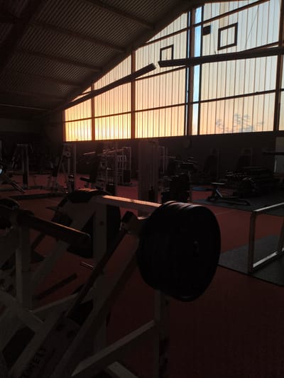 The image depicts the interior of a dimly lit gym during sunset. The gym equipment, including weight machines and barbells, are silhouetted against the warm orange and blue hues of the setting sun visible through a large, corrugated window spanning the length of the building. The floor is covered with red and black mats. The scene is devoid of people, creating a sense of stillness and quiet. Based on the provided information, this gym is located in Pegnitz, Germany.