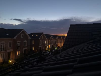 This image depicts a twilight scene in Baranovo, Russia, showing a row of townhouses with lights on in their windows. The foreground is dominated by the dark, tiled roof of a building, casting it into shadow. The sky above is a gradient of pale blue and grey, with clouds present, and a faint orange glow suggesting the setting sun or the dawn. Several cars are parked in front of the houses. The architecture appears to be European-style, with brick facades and multi-paned windows. No people are visible in the image.