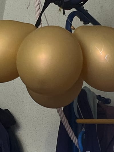 The image shows a close-up of a cluster of gold balloons. The balloons are round and appear to be made of a metallic material, reflecting light. One of the balloons has faint white markings that resemble letters or symbols. The balloons are arranged vertically, with some overlapping each other. In the background, a blue railing or play structure is visible, along with a thick rope that has a pinkish hue. Part of a yellow pole is also present. The overall setting seems to be indoors, possibly at a party or event, given the balloons. The lighting suggests it could be daytime, but it's difficult to determine the exact time. There are no people clearly visible in the frame, only the objects. The textures in the background are a bit grainy, possibly indicating a wall with a textured finish. No specific location cues pinpoint Muravlenko, Russia, other than the provided context.