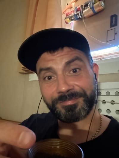 A close-up, selfie-style shot shows a man in a black baseball cap and a black t-shirt. He has a beard and is wearing earbuds. He is holding a small, dark-colored cup. Behind him, several cans are taped together and attached to a cardboard surface. A curtain is visible to the left, and a window with a white frame and horizontal blinds is to the right. The overall impression is that of an indoor, possibly cramped, living or working space, with no clear indicators of a specific location in Pevek, Russia, beyond the provided context.